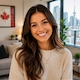 Smiling young woman with long dark hair in a modern apartment with city skyline windows and a framed Canadian flag artwork in the background.